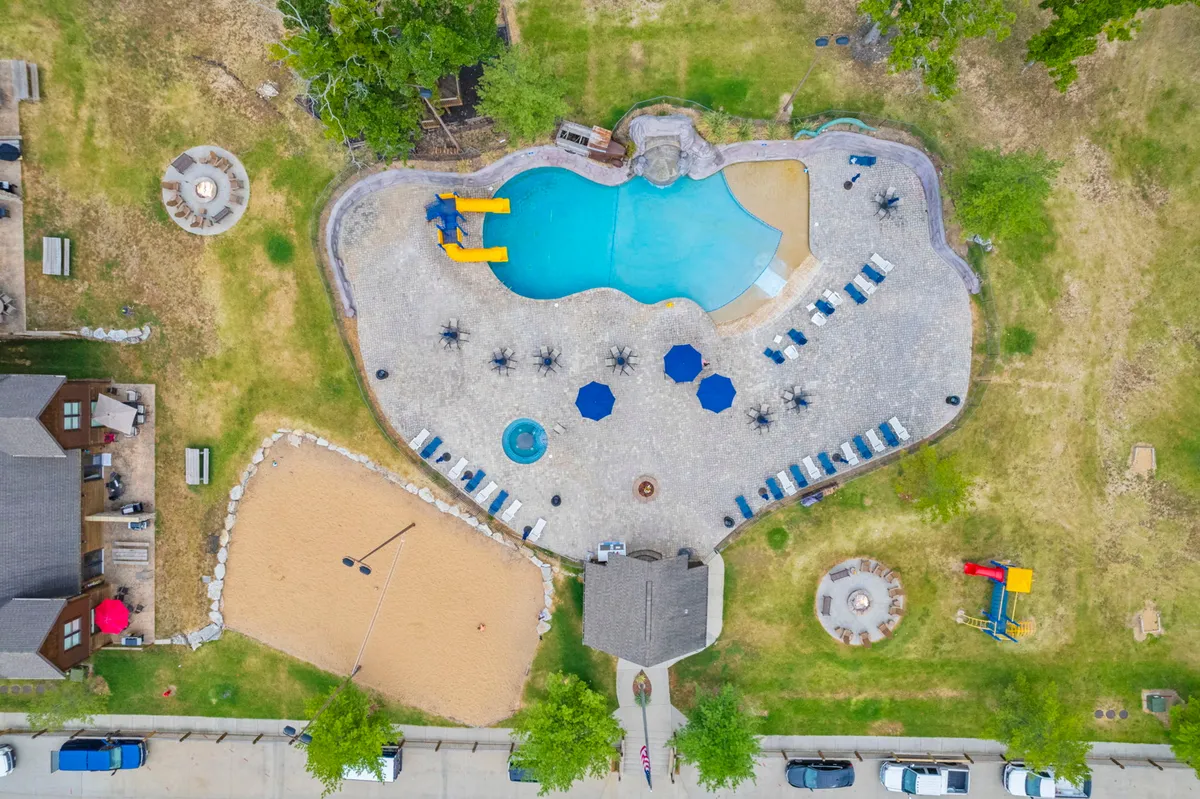 A lively exterior view featuring a resort-style swimming pool with a water slide, adjacent hot tub, beach volleyball area, and rows of lounge chairs. The area is framed by mature trees and open skies, creating the perfect setting for family fun and relaxation.