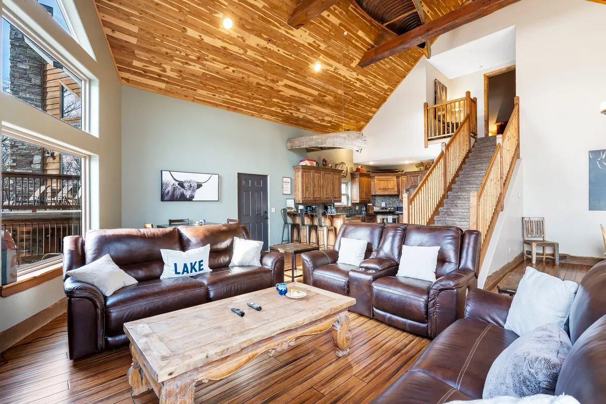 The image showcases a cozy living room with rustic wooden ceilings, large windows that frame beautiful sunrise views, comfortable leather sofas, and a wooden coffee table—creating a perfect space for gathering and relaxation.