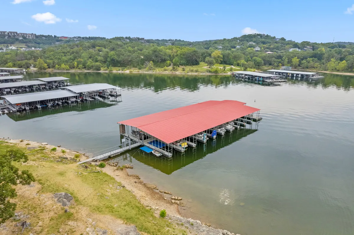 A tranquil lakeside scene featuring a red-roofed dock extending over calm water, surrounded by lush greenery and boats moored along the edge. The setting invites boating, fishing, swimming, and relaxing at the water's edge.
