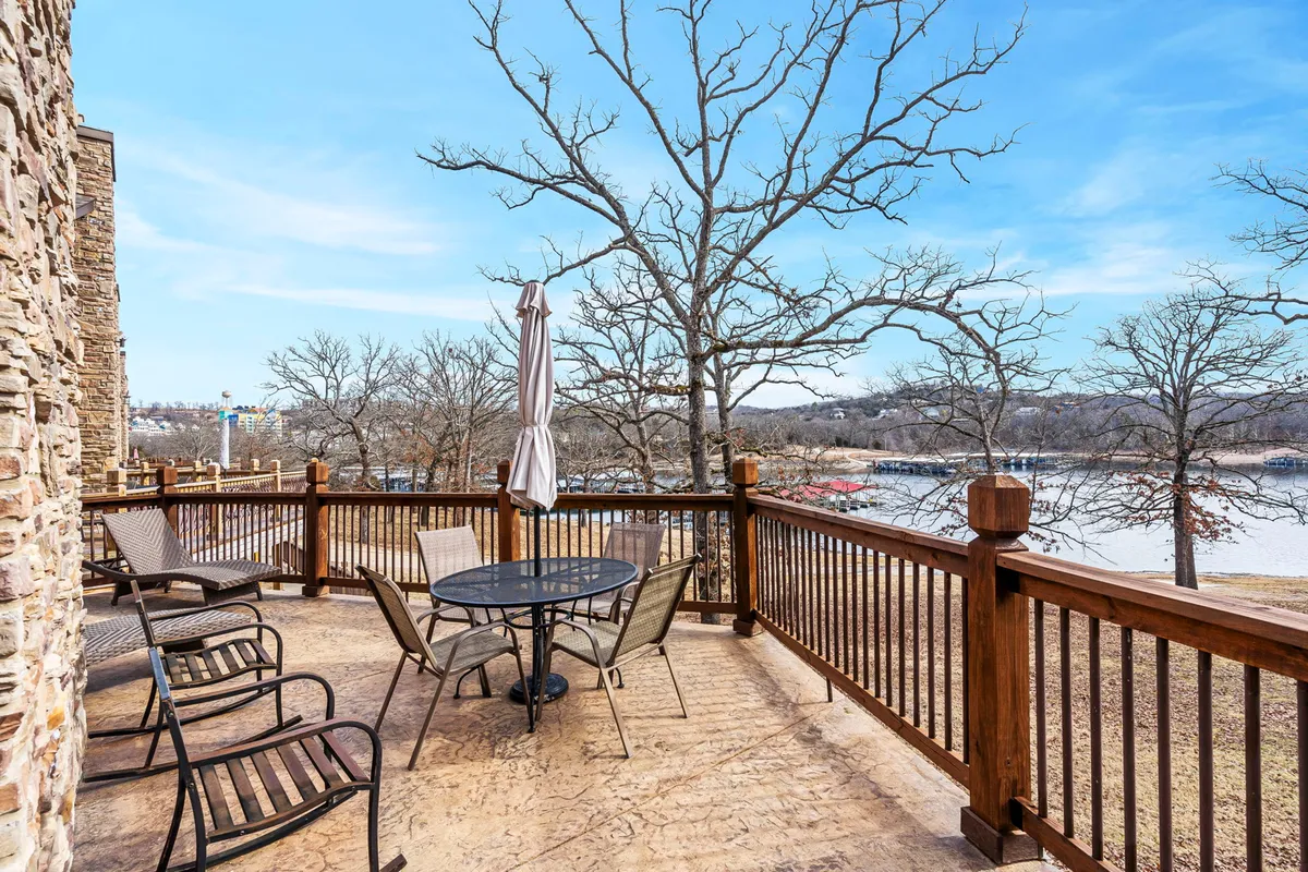 This image captures a serene patio area furnished with comfortable outdoor furniture, a parasol, and a direct view of the lake in the background. The scene exudes tranquility, with bare trees hinting at both privacy and natural beauty. The layout suggests ample space for dining, relaxing, or gathering with friends and family while enjoying the lakeside atmosphere.