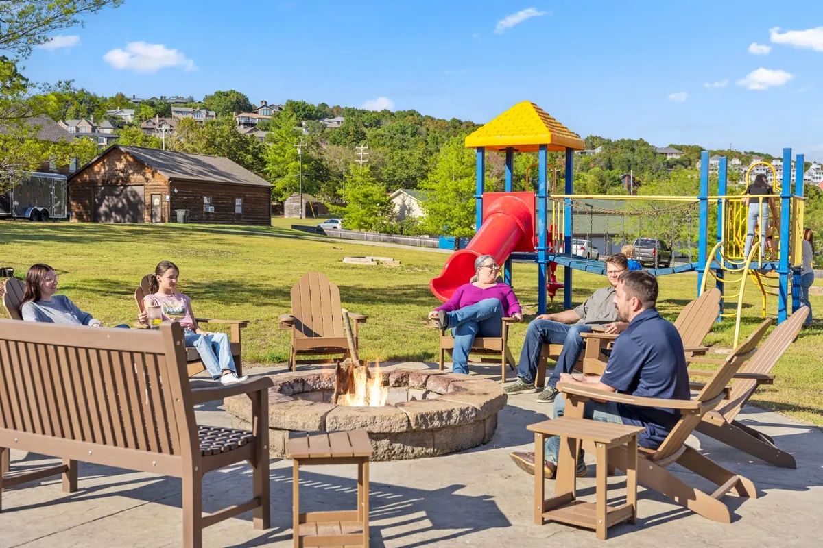 An inviting exterior gathering area with a fire pit surrounded by seating, adjacent to a playground. Children are seen playing while adults gather around the fire, suggesting an evening of s'mores, laughter, and storytelling.