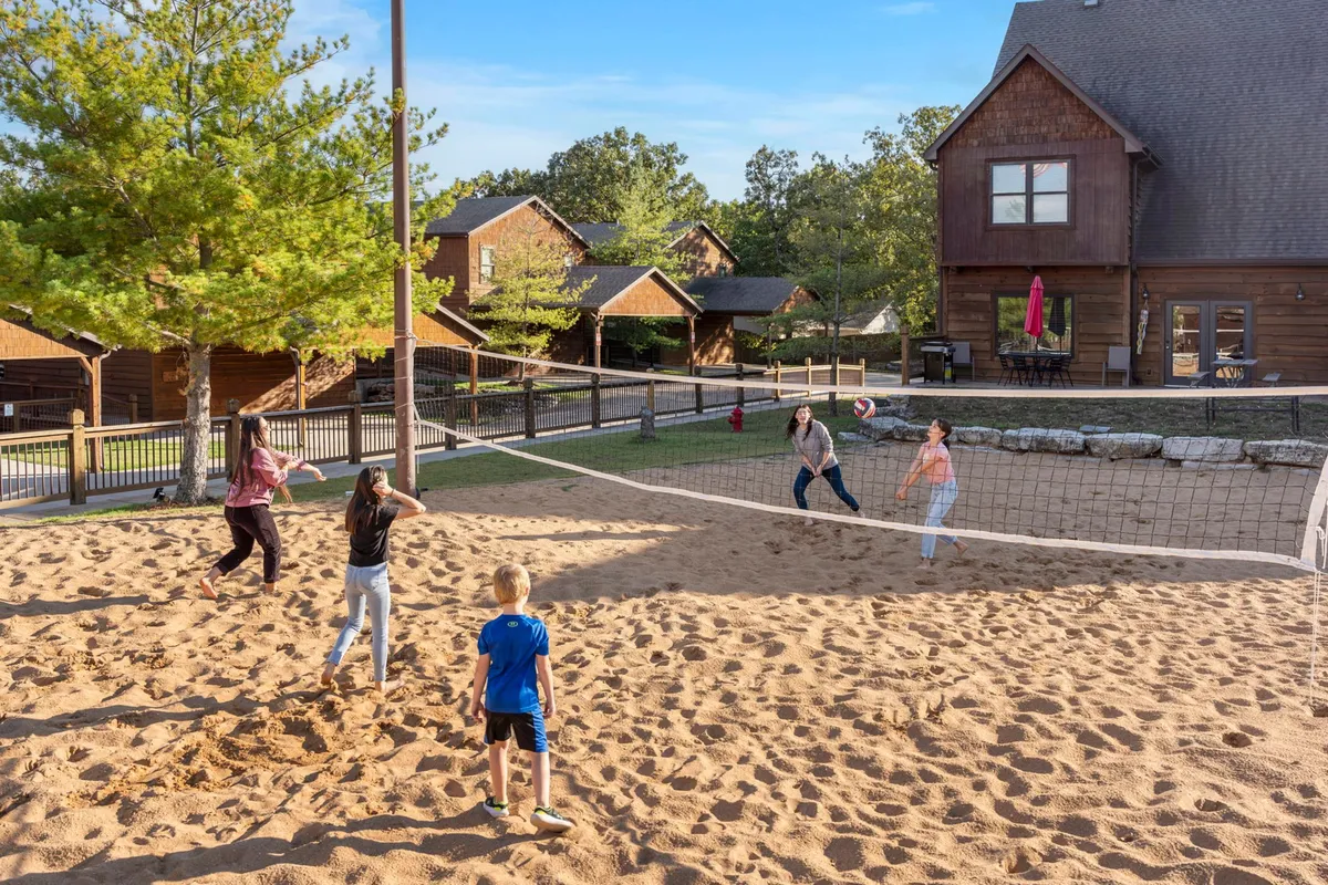 A dynamic scene of guests playing sand volleyball on a court surrounded by green trees and cozy resort cabins. The joyful energy is palpable as people engage in friendly competition, laughter, and outdoor bonding.