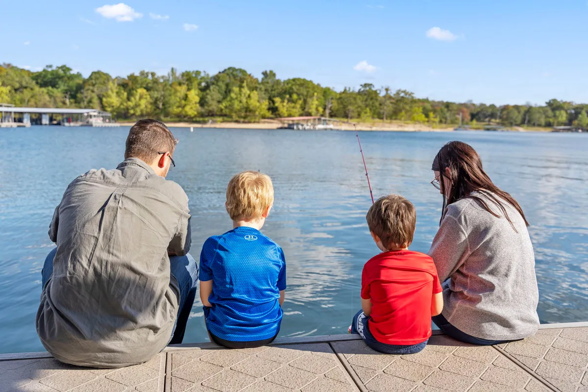 A family enjoys fishing on the calm lakeshore, with tall trees in the background adding to the peaceful, natural setting. Smiles and laughter highlight the joy of family bonding and experiencing the outdoors together.